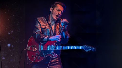 Ein Musiker in Lederkleidung singt leidenschaftlich ins Mikrofon, während er auf der Bühne bei dunkelblauer Beleuchtung E-Gitarre spielt.