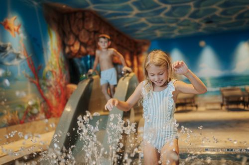 Ein Mädchen spielt im Wasser vor einer kleinen Rutsche, auf der ein Junge steht. Wandmalereien mit Fischen und Korallen gestalten den Kinderbereich der IKUNA Spa- & Wellnesswelt.