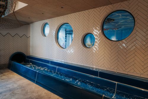Auslauf der LED-Röhrenrutsche mit blauem Auffangbecken neben einer hell gefliesten Wand mit runden Fenstern zum Kinderbereich der IKUNA Spa- & Wellnesswelt.