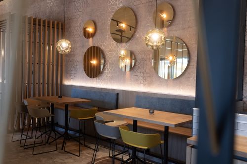IKUNA Erlebnisbad Bistro Moderner Restaurantbereich mit Holztischen, bunten Stühlen, blauen Sitzbänken, runden Spiegeln, gemusterten Wänden und warmen Pendelleuchten.