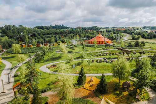 IKUNA Naturresort Eine malerische Luftaufnahme eines üppig grünen Parks mit gewundenen Wegen, Dinosaurierstatuen und einem leuchtend orangefarbenen Zirkuszelt im IKUNA Naturresort. Der Himmel ist teilweise bewölkt.