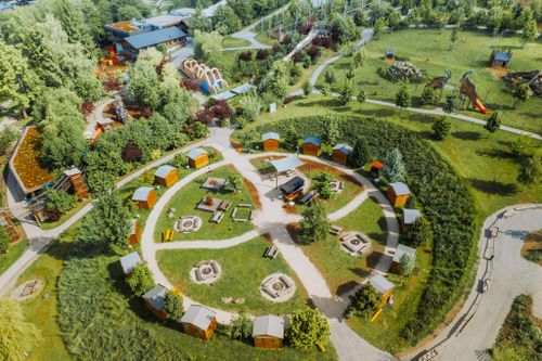 IKUNA Naturresort Letecký pohled na přírodní park IKUNA s kruhem malých chatek obklopených turistickými stezkami. Lavičky uprostřed a ohniště navozují atmosféru kempu. V blízkosti dodávají areálu hravý nádech velké skluzavky a hřiště.
