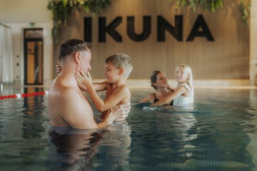 Zwei Erwachsene halten jeweils ein Kind im Indoorpool der IKUNA Spa- & Wellnesswelt. Im Hintergrund ist der IKUNA-Schriftzug an einer Holzwand zu sehen.
