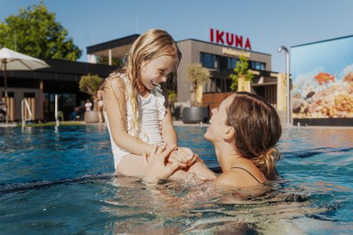 Eine Mama mit ihrer Tochter, die im sonnigen IKUNA Erlebnisbad fröhlich lächeln, im Hintergrund eine sommerliche Kulisse mit Umrissen von Gebäuden, wunderschönen Palmen und ausreichend Platz zum Entspannen.