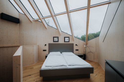 Bett steht zentral im modernen Schlafzimmer mit Holzwänden. Oberlichtfenster lassen Tageslicht herein. Pflanzen und minimalistische Dekoration ergänzen das Ambiente.