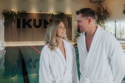 Eine Frau und ein Mann in weißen Bademänteln lächeln sich am Indoorpool der IKUNA Spa- und Wellnesswelt an. Im Hintergrund sind Pflanzen und der IKUNA-Schriftzug an einer Holzwand zu sehen.