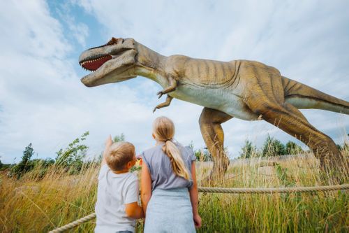 IKUNA Naturresort Kinder blicken zu einer großen Dinosaurierskulptur auf einem offenen Feld, im IKUNA Naturresort, hinauf. Der Junge zeigt aufgeregt darauf, und das Mädchen steht neben ihm. Die Szene wirkt lebhaft und weckt die Neugier.
