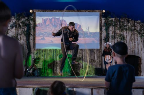IKUNA Zirkusshow Vor einer Wüstenkulisse wirbelt ein Artist gekonnt ein Lasso, während er auf der Bühne herumhüpft. Ein begeistertes Publikum schaut zu und sorgt für eine lebhafte Atmosphäre bei der IKUNA-Zirkusshow.
