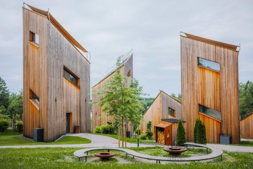 IKUNA Naturresort Moderní dřevěné budovy se šikmými střechami stojí uprostřed bujné zeleně pod zamračenou oblohou. Zakřivené cestičky a kruhové lavičky dotvářejí klidnou atmosféru resortu IKUNA Naturresort.