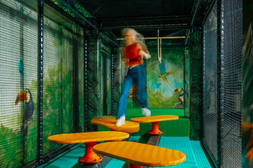 IKUNA Naturresort Eine Person, die sich in Bewegung befindet, erscheint verschwommen, während sie auf lebhaften, runden Plattformen im Spielbereich, des IKUNA Ninja-Dschungels, mit Dschungelmotiven herumhüpft. Energiegeladen, farbenfroh, mit Tukanen auf üppigen Wandmalereien.