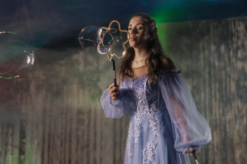 Frau in langem, fliederfarbenem Kleid bläst Seifenblasen mit Blasenstab. Der Hintergrund ist verschwommen, bunte Beleuchtung erzeugt zauberhafte Atmosphäre.