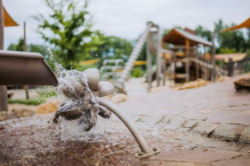 IKUNA Naturresort Venkovní hřiště s vodním prvkem, z něhož stříká voda z rotujícího prvku. V pozadí jsou vidět dřevěné konstrukce a zeleň v resortu IKUNA Naturresort.