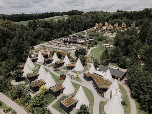 Luftaufnahme der IKUNA-Unterkünfte mit Tipis, Chalets und Campingplatz in grüner, bewaldeter Umgebung.