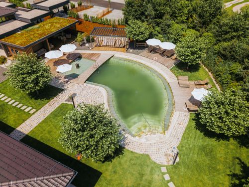 IKUNA Naturresort Pohled z ptačí perspektivy na klidný přírodní bazén obklopený bujnou zelení a dřevěnou terasou. Bílé slunečníky a lehátka dotvářejí v resortu IKUNA Naturresort klidnou a příjemnou atmosféru.
