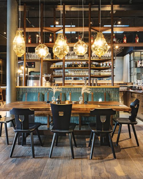 Gemütliches Restaurantinterieur mit warmer Beleuchtung, einem Holztisch und schwarzen Stühlen. Im Hintergrund sind auf Regalen Flaschen und Gläser ausgestellt.