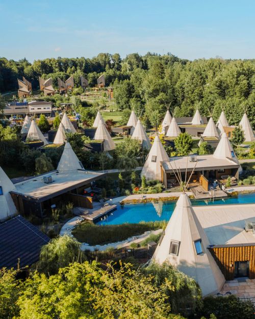 Luftaufnahme von zahlreichen Hütten im Tipi-Stil, umgeben von üppigem Grün und einem Naturschwimmteich unter strahlend blauem Himmel.