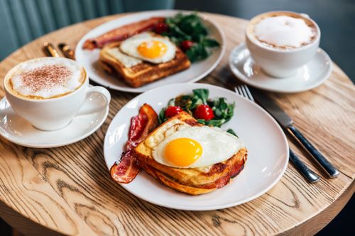 Frühstück mit geröstetem Brot, Spiegeleiern, Speck, Salat und Kirschtomaten, serviert mit Cappuccino auf einem Holztisch.