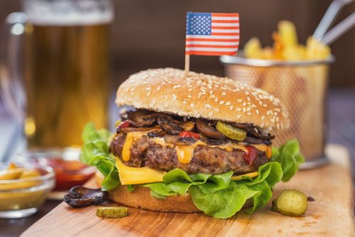 IKUNA Naturresort Ein saftiger Cheeseburger mit Salat, Gewürzgurken, Champignons und einem Zahnstocher in Form einer amerikanischen Flagge auf einem Holzbrett im IKUNA Naturresort. Im Hintergrund: Pommes frites in einem Korb und ein Glas Bier.