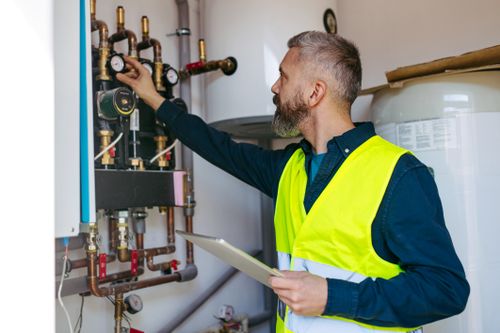Stellenausschreibung Installateur bei IKUNA Ein Wartungsmitarbeiter in einer Warnweste überprüft im Technikraum bei IKUNA Naturresort industrielle Rohrleitungen und Messgeräte und stellt dabei ein Ventil ein, während er ein Tablet in der Hand hält.