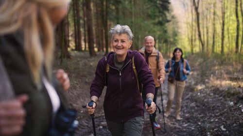 Eine Gruppe von vier Erwachsenen wandert gemeinsam auf einem Waldweg. Im Mittelpunkt geht eine ältere Frau lächelnd mit Wanderstöcken, während die anderen Personen hinter ihr folgen.