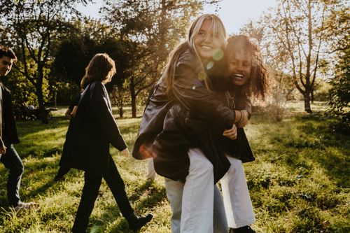 Zwei Freunde lachen gemeinsam in einem sonnigen Park, während eine Person die andere auf dem Rücken trägt. Im Hintergrund spazieren weitere Personen zwischen Bäumen und über eine Wiese.