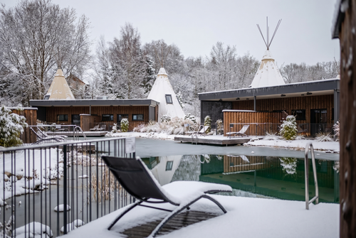 IKUNA Lake Side Tipi Suiten in WInterlandschaft Schneebedeckte Tipis und moderne Hütten umgeben einen stillen, teilweise zugefrorenen Teich. Die kahlen Bäume im Hintergrund unterstreichen die friedliche, winterliche Szenerie.
