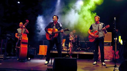 Eine Band spielt voller Energie auf der Bühne, beleuchtet von stimmungsvollem grünem und gelbem Licht. Der Leadsänger spielt Akustikgitarre, flankiert von einem Bassisten und einem Gitarristen. Der Schlagzeuger steht im Hintergrund. Alle tragen schwarz-weiß gestreifte Hemden und wirken engagiert und leidenschaftlich.