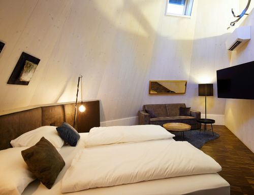 Lake Side Tipi Suite Gemütliche IKUNA Lake Side Tipi Suite mit Doppelbett, weißer Bettwäsche, Sofa, Fernseher und heller Holzverkleidung unter hoher Decke.