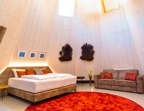Wellness Tipi Suite Geräumige IKUNA Wellness Tipi Suite mit gemütlichem Doppelbett, Sofa, rotem Teppich und heller Holzverkleidung unter hoher Decke.