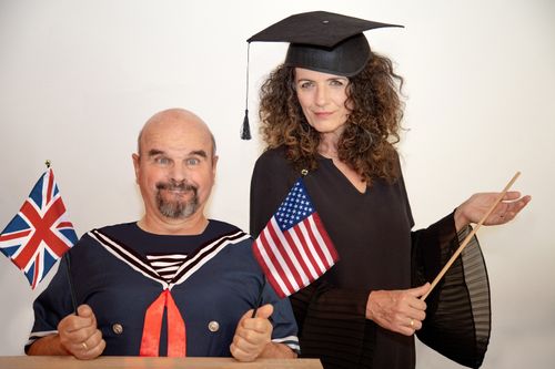 Ein Mann in einer Matrosenuniform hält eine britische Flagge in der Hand, während eine Frau mit einer Abschlusskappe eine US-Flagge und einen Zeigestock hält; beide lächeln.