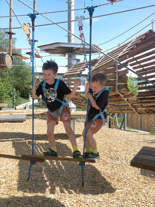 Zwei kleine Jungen in Klettergurten überqueren einen hölzernen Schwebebalken auf einem Seilgarten im Freien. Unter dem strahlend blauen Himmel wirken sie konzentriert und abenteuerlustig.