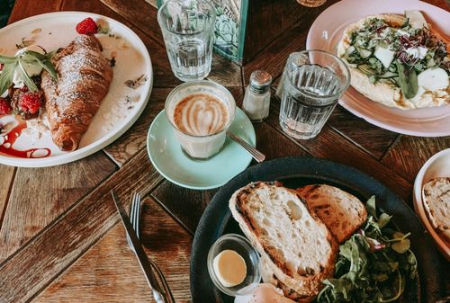 Frühstücksteller mit Croissant, Brot, Salat und Kaffee auf Holztisch, umgeben von Gläsern und Besteck in einem Café-Setting.