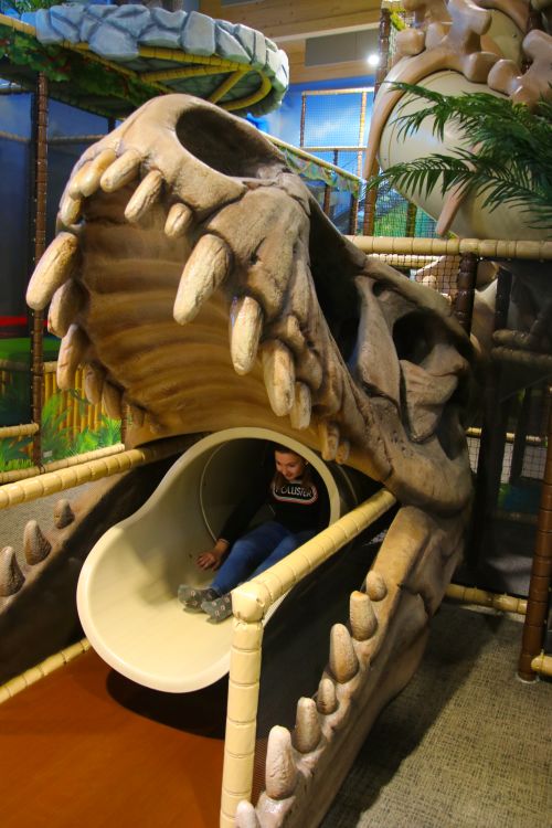 IKUNA Naturresort In dem Indoor Spielplatz, KIDS WORLD, im IKUNA Naturresort rutscht ein Kind aus dem offenen Maul eines Dinosauriers heraus. Die Kulisse ist farbenfroh und mit Dschungel-Motiven gestaltet, was ein Gefühl von Abenteuer vermittelt.
