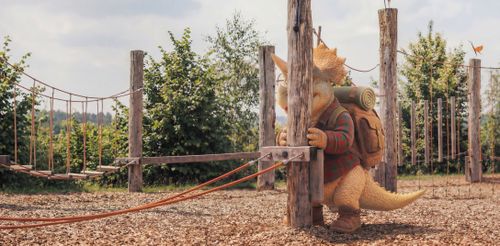 Ein Dinosaurier in Wanderkleidung lehnt schüchtern an einem Holzpfosten auf einem Spielplatz im Freien. Die sonnige und grüne Umgebung sorgt für eine verspielte Stimmung.