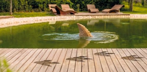 Ein ruhiger Teich, aus dem eine verspielte Dinoflosse aus dem Wasser ragt. Auf der Holzterrasse sind Dinofußstapfen zu sehen, vor dem Hintergrund Wellnessoase und Liegestühlen.