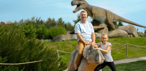 Ein Mann und ein junges Mädchen sitzen lächelnd auf einer Dinosaurierstatue in einem Park, im Hintergrund ist ein großes Dinosauriermodell zu sehen.