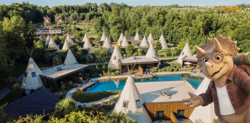 IKUNĚ Přírodní resort Letecký pohled na malebné, zalesněné letovisko s trojúhelníkovými chatkami a centrálním bazénem. Vpravo stojí s úsměvem na tváři přátelská kreslená postavička dinosaura.
