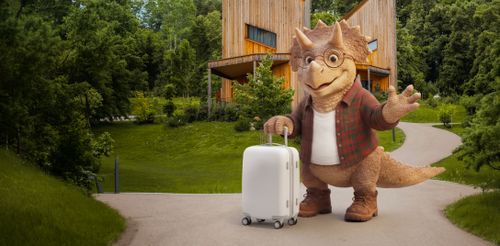 Ein Cartoon-Triceratops mit Brille und Flanellhemd winkt fröhlich, während er mit einem Koffer auf einem Weg steht. Im Hintergrund sind ein modernes Holzgebäude und üppiges Grün zu sehen.
