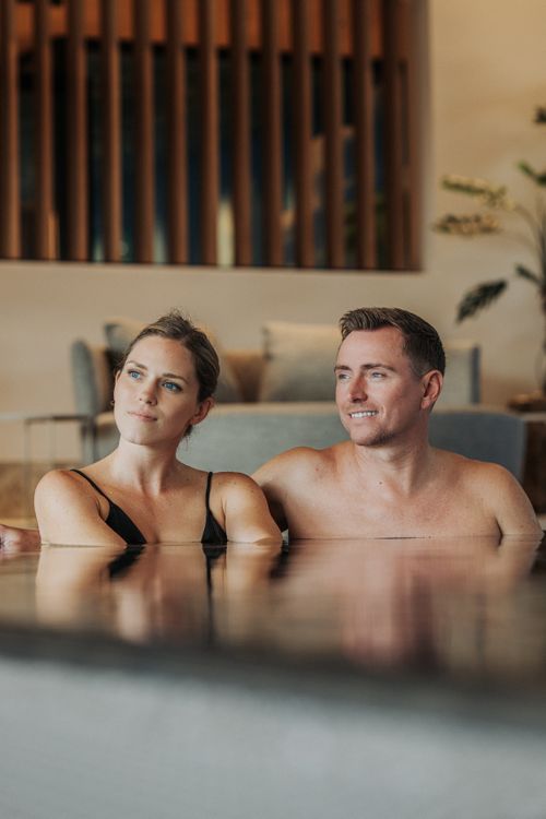 Ein Paar entspannt gemeinsam im Indoorpool der IKUNA Spa- & Wellnesswelt in Oberösterreich. Hinter ihnen sind gepolsterte Ruheplätze und eine stilvolle Holzwand zu sehen.