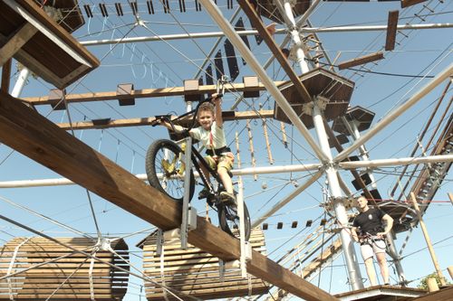 Kind fährt Fahrrad über Holzplanke in Kletterparcours, umgeben von Seilen und Türmen unter klarem Himmel.