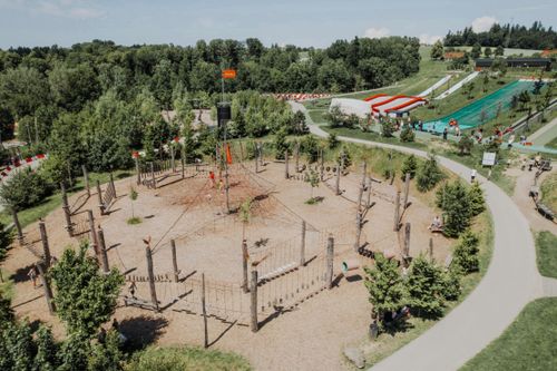 Kletterelement im Erlebnispark Kletterpark mit Seilen und Plattformen, umgeben von Bäumen. Besucher nutzen die Kletterelemente. Im Hintergrund eine Rutsche und Spazierwege.