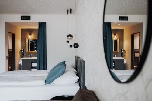 IKUNA Naturresort Moderní hotelový pokoj, který se odráží v kulatém zrcadle, s úhledně ustlanou postelí s tyrkysovými polštáři, závěsnými svítidly a koupelnou s tyrkysovými závěsy a útulnou atmosférou v IKUNA Naturresort.