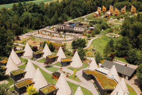 IKUNA Naturreosrt Luftaufnahme des IKUNA Naturresort mit zahlreichen weißen Tipis in einer ruhigen, idyllischen Umgebung, die sich durch ihre grünen Wiesen, gewundenen Wege und Holzhütten auszeichnet.
