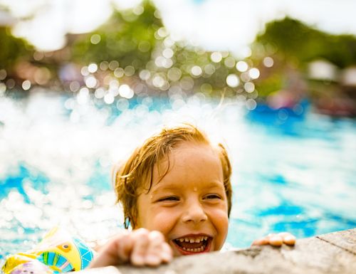 Kind schwimmt im Becken und schaut über den Beckenrand in die Kamera.