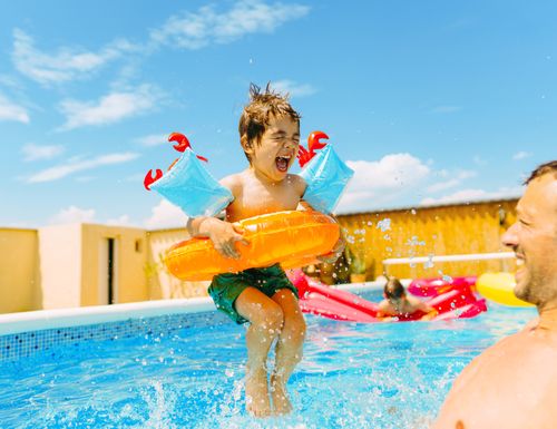 Kind hüpft mit Schwimmflügerl und Schwimmreifen in den Pool wo Papa wartet