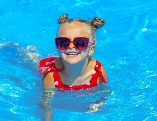 Kleines Mädchen chillt im Knietiefen Wasser mit rotem Badeanzug und Sonnenbrille von Mama.