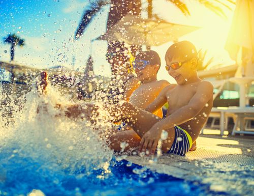 IKUNA_Erlebnisbad_Kinder_Poolrand Zwei Jungs sitzen am Beckenrand und spielen mit ihrem Beinen im Wasser, dass es spritzt