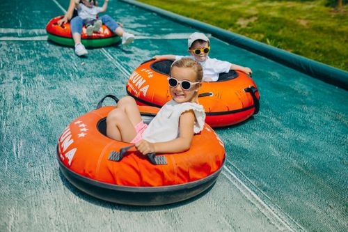 IKUNA Naturresort Kinder mit Sonnenbrillen lächeln, während sie auf bunten Schlauchbooten eine Rutsche hinunterrutschen. Die Szene vermittelt Freude und Aufregung an einem sonnigen Tag im IKUNA Naturresort.