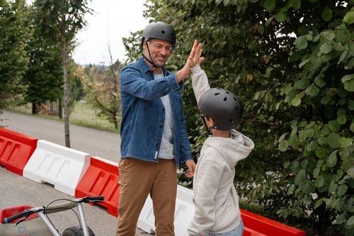 Ein Mann und ein Kind, beide mit Helmen, geben sich im Freien lächelnd ein High Five. Der Mann trägt eine Jeansjacke, das Kind einen grauen Kapuzenpulli; die Szene strahlt Freude und Teamgeist aus.