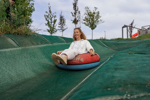 Eine Frau in weißer Kleidung rutscht fröhlich auf einem aufblasbaren Reifen eine grüne Kunststoffrutsche an einem Hang hinunter, im Hintergrund sind einige Bäume und ein bewölkter Himmel zu sehen.